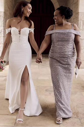 Two women walking hand-in-hand, one in a white wedding dress and the other in a silver evening gown, with a stone building in the background.
