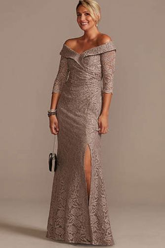 Woman wearing a long, off-shoulder lace dress with a slit on a plain background