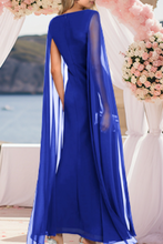 Load image into Gallery viewer, Woman wearing a blue dress with a cape in front of floral decorations.