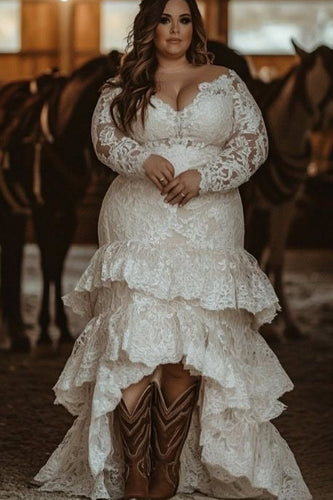 Woman in a lace wedding dress with cowboy boots standing in a barn with horses.